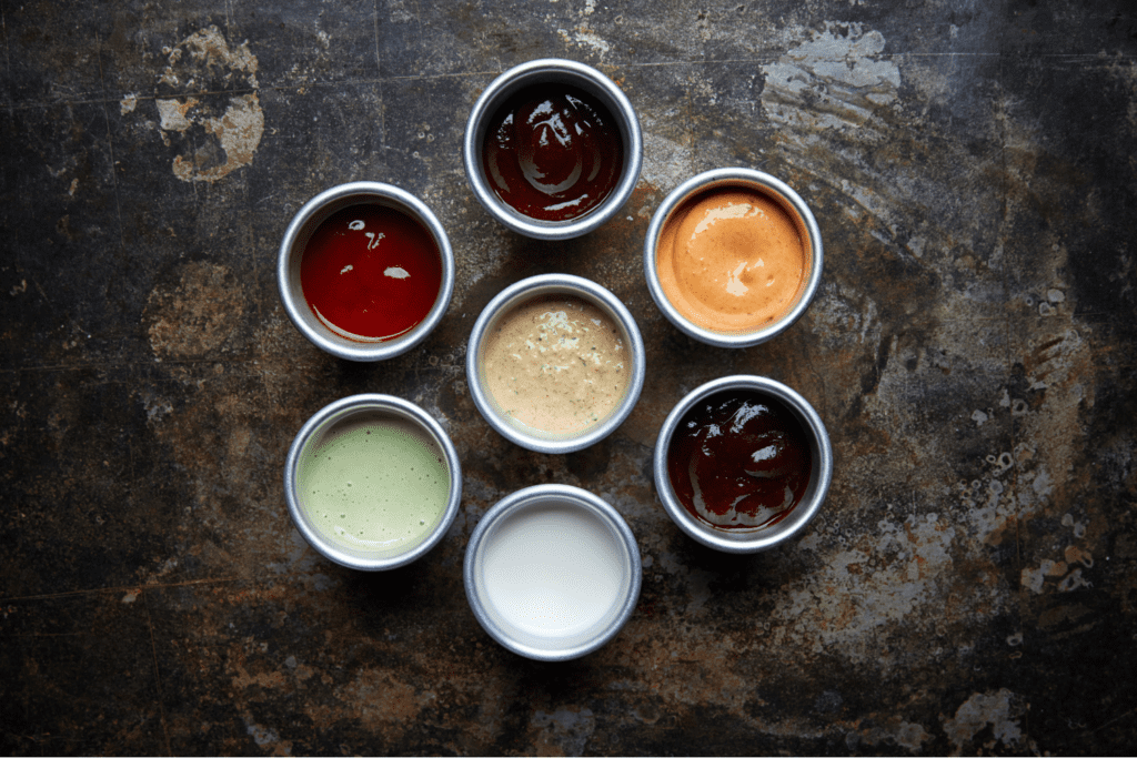 Seven different dipping sauces in white ramekins against a dark background